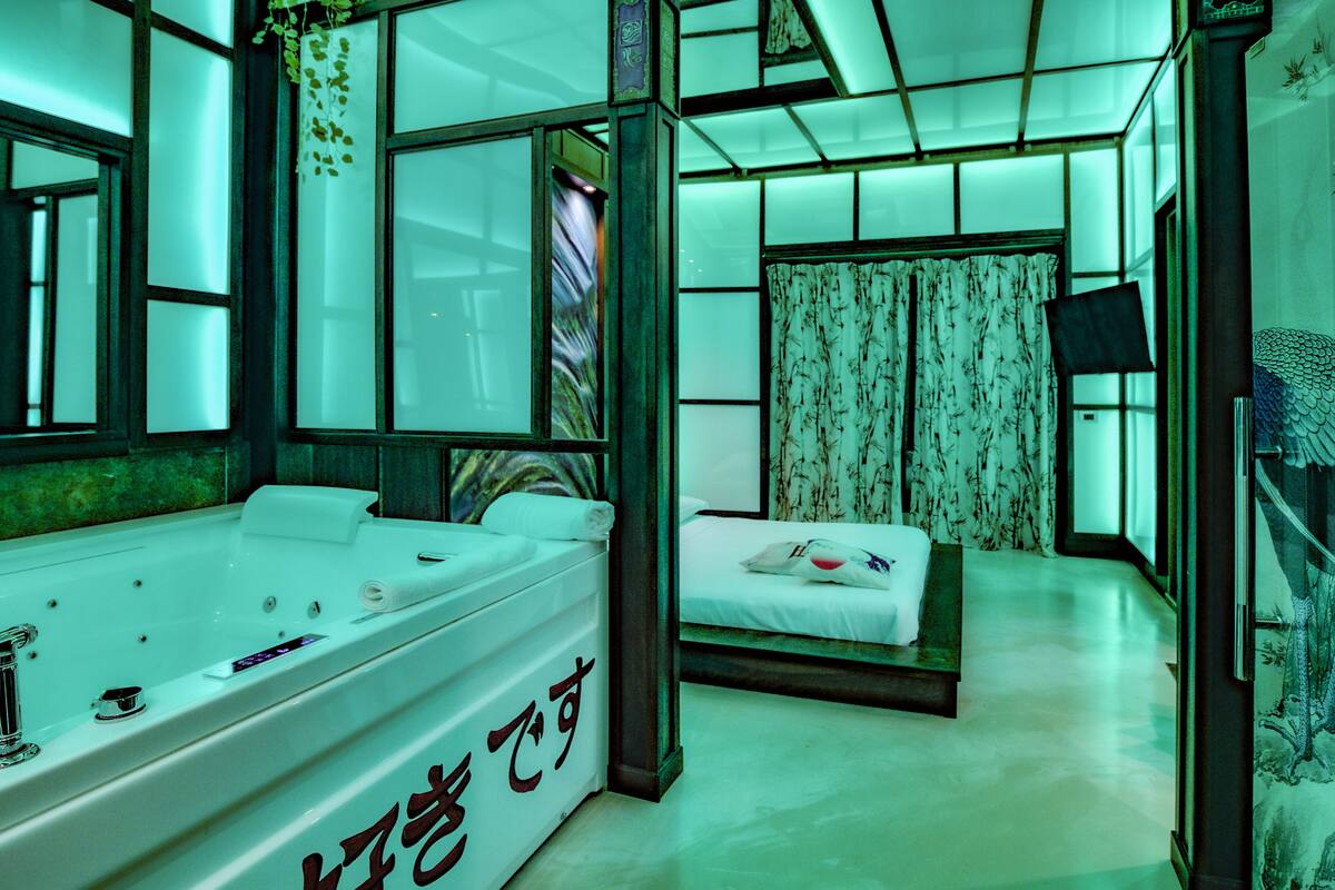 A modern hotel room with a large bathtub, a bed with a white bedspread, and a traditional-style window with curtains.