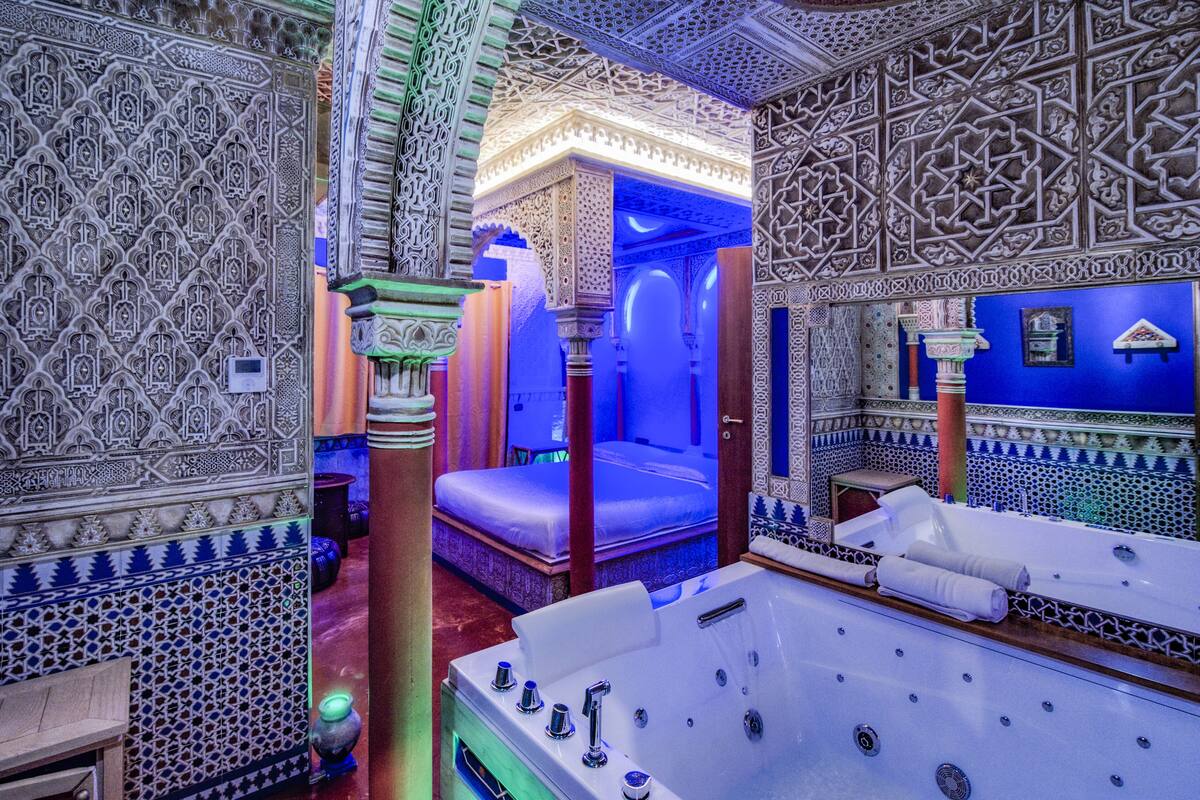 A luxurious bathroom with intricate tile work, a large bathtub, and a bed with a blue-lit canopy.