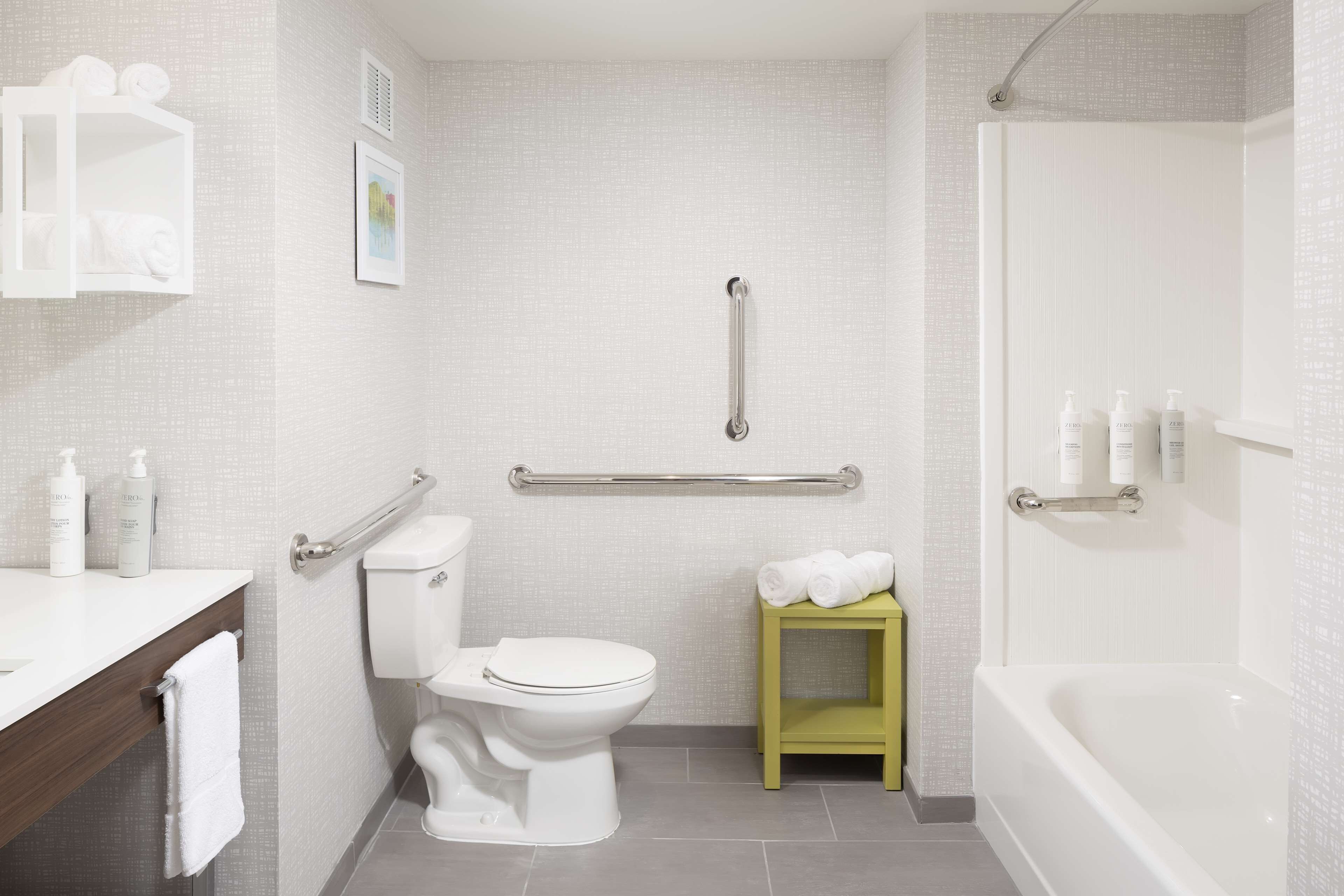 A bathroom with a toilet, a small green stool with towels, a handrail, and a bathtub.