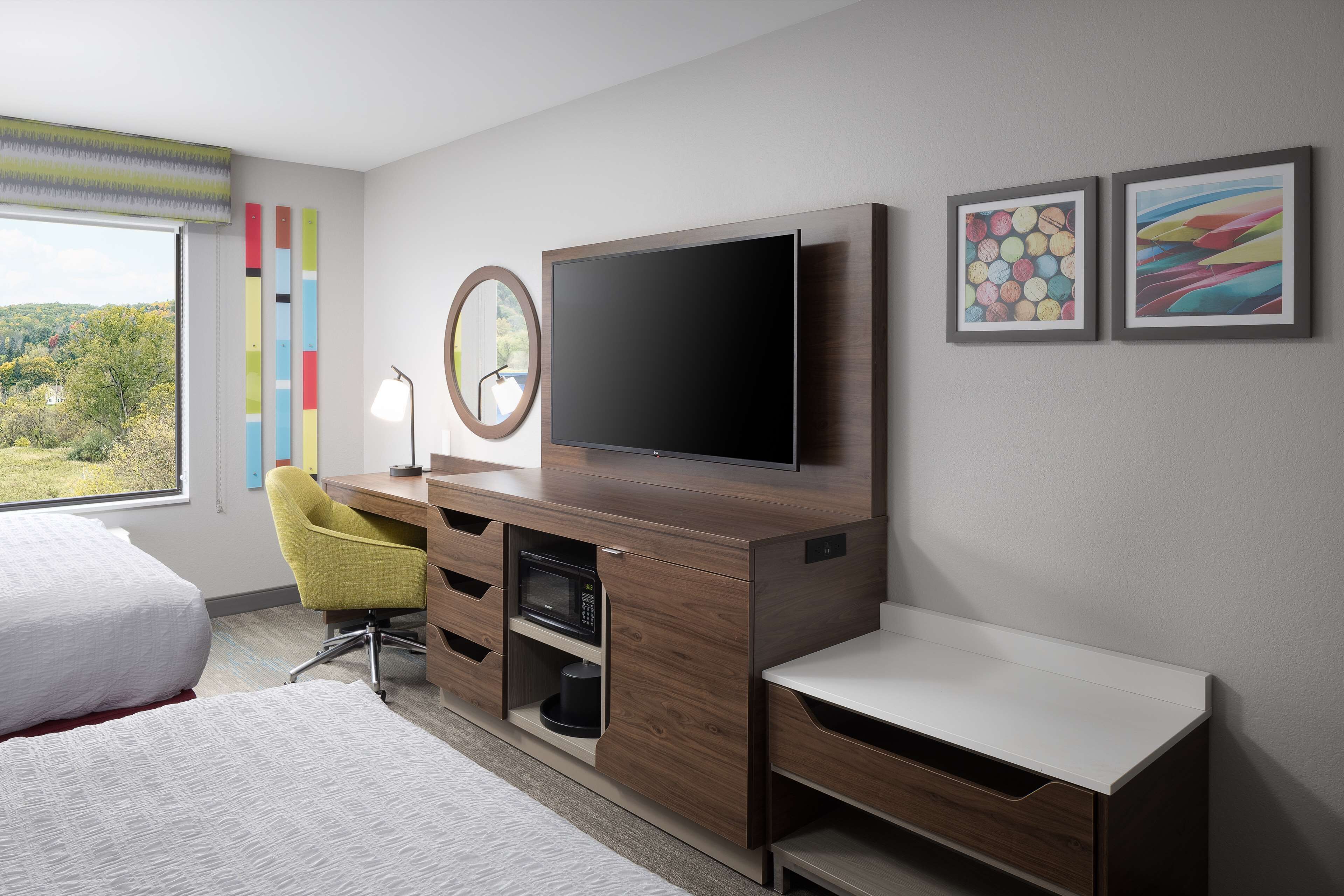 A modern hotel room with a large bed, a desk with a flat-screen TV, a yellow chair, and a view of the outdoors.