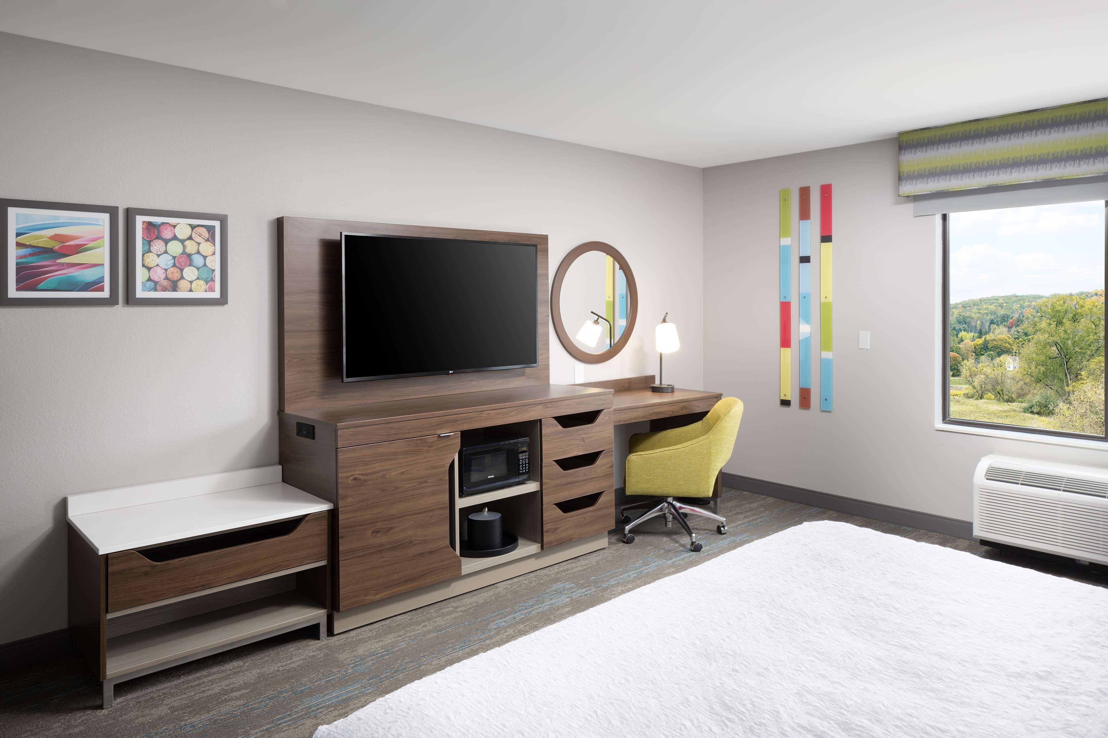 A hotel room with a bed, a desk with a flat-screen TV, a microwave, a chair, and a large window with a view of trees.