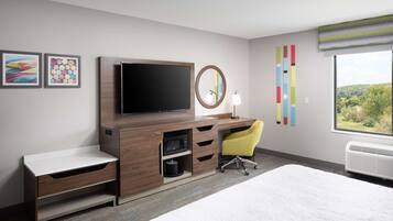 A hotel room with a bed, a desk with a flat-screen TV, a microwave, a chair, and a large window with a view of trees.