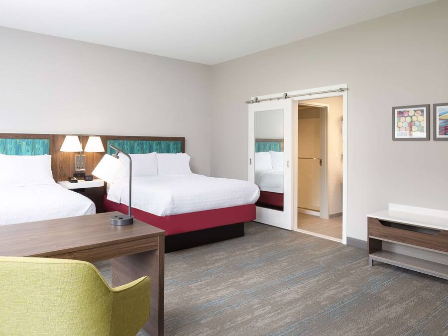 A hotel room with two beds, a desk, a chair, a bedside table with lamps, a wardrobe, and a small bedside table.