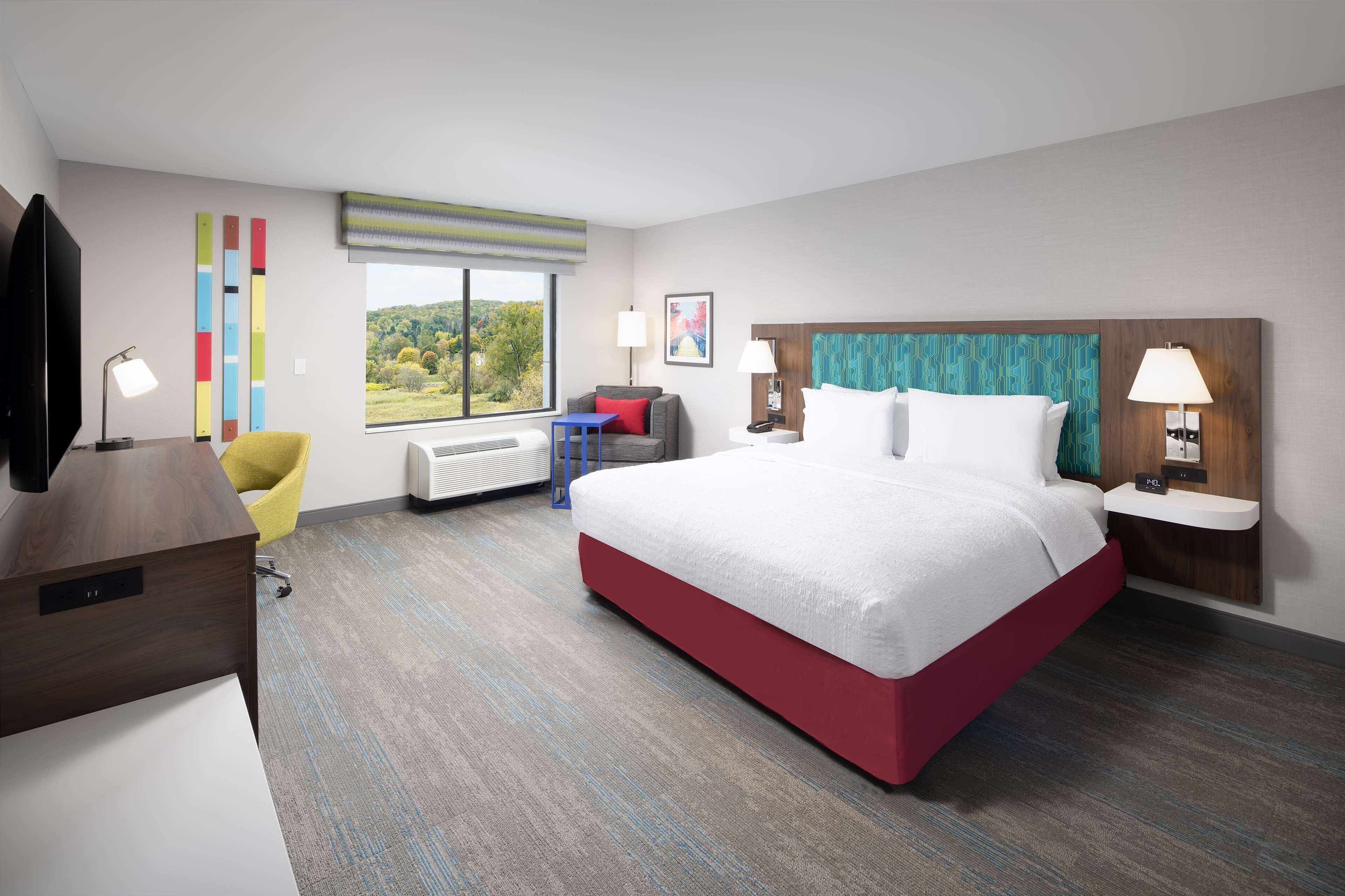 A hotel room with a large bed, a desk, a chair, a TV, a window with a view of greenery, and a wall with colorful vertical elements.