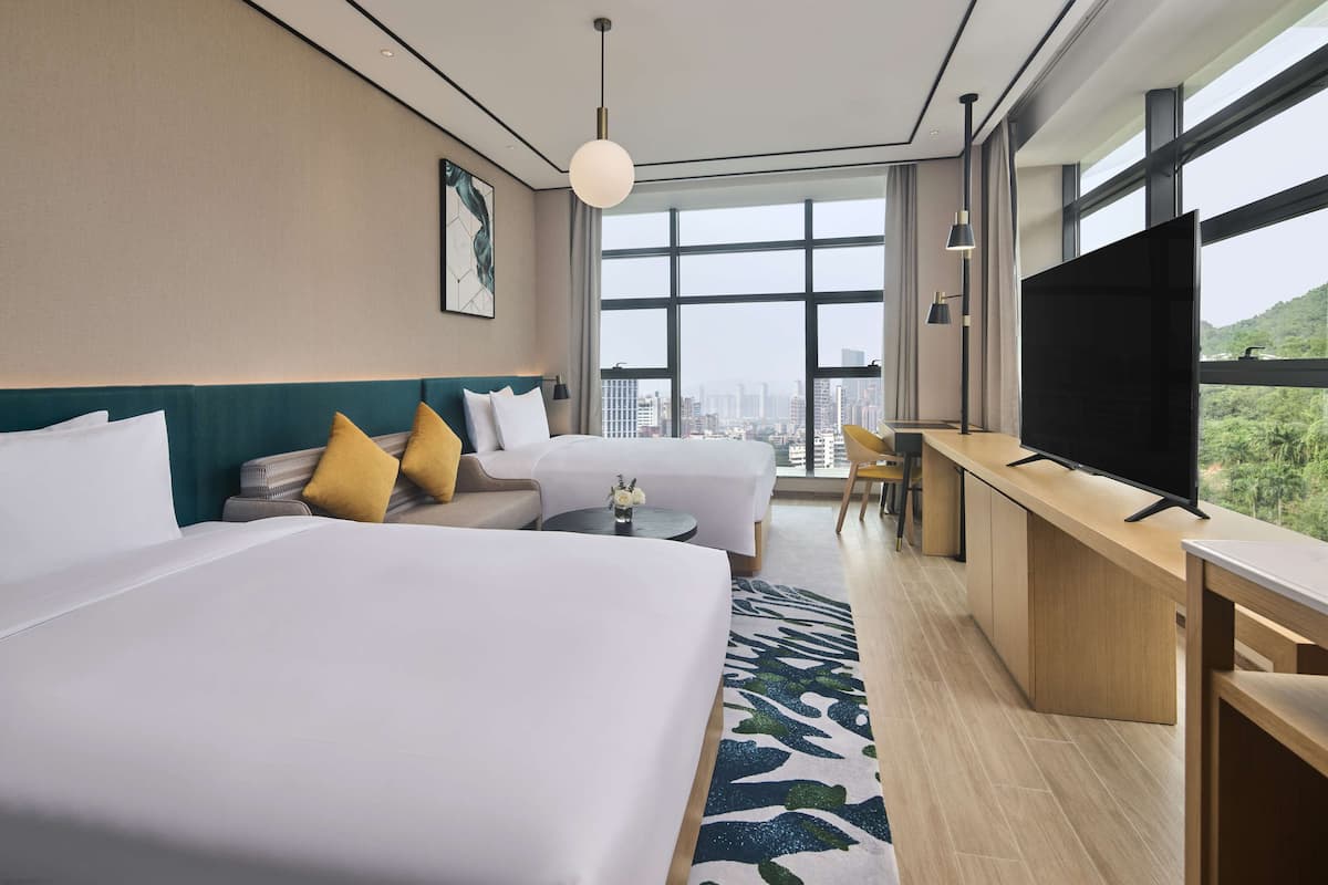A hotel room with two beds, a desk, a TV, and a large window with a city view.
