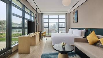 A modern hotel room with a large window, a bed, a sofa, a desk, and a television.