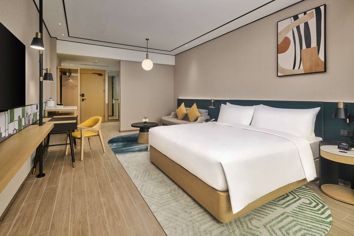 A modern hotel room with a large bed, a desk with a chair, a small table, and a wall-mounted TV.