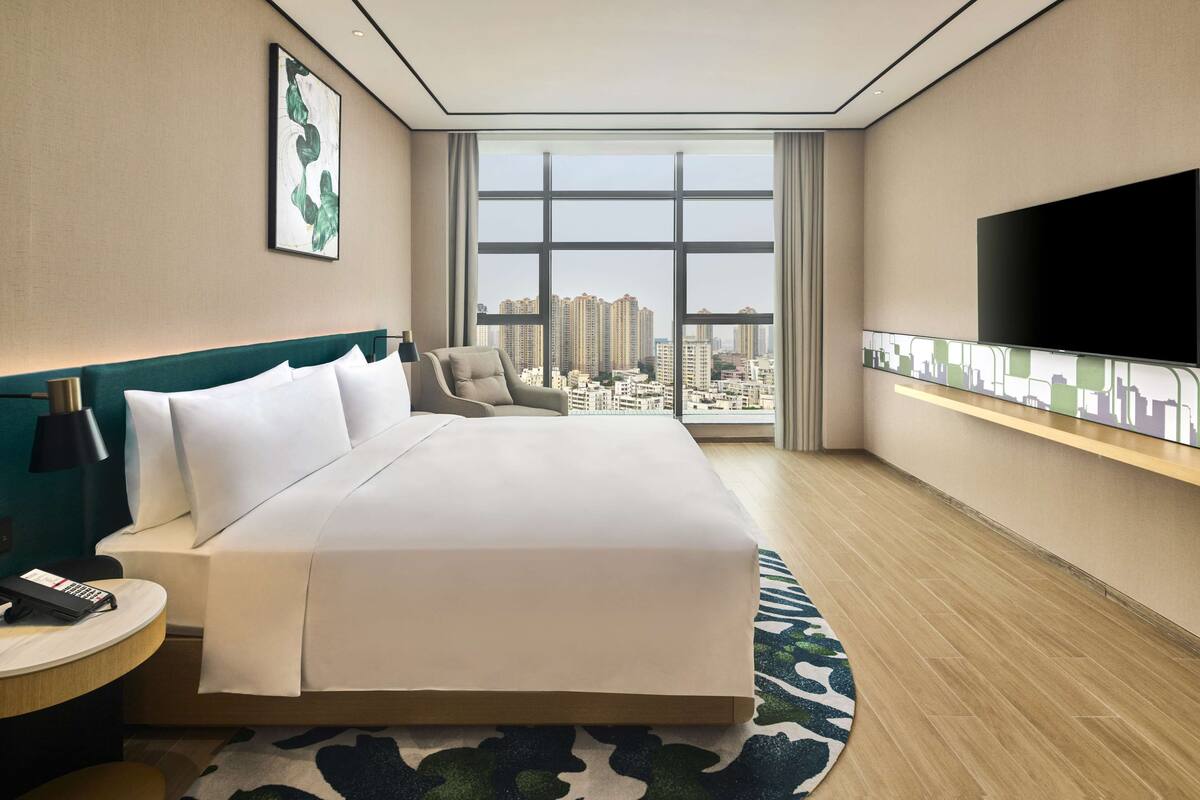 A hotel room with a large bed, a TV mounted on the wall, and a view of the city through large windows.