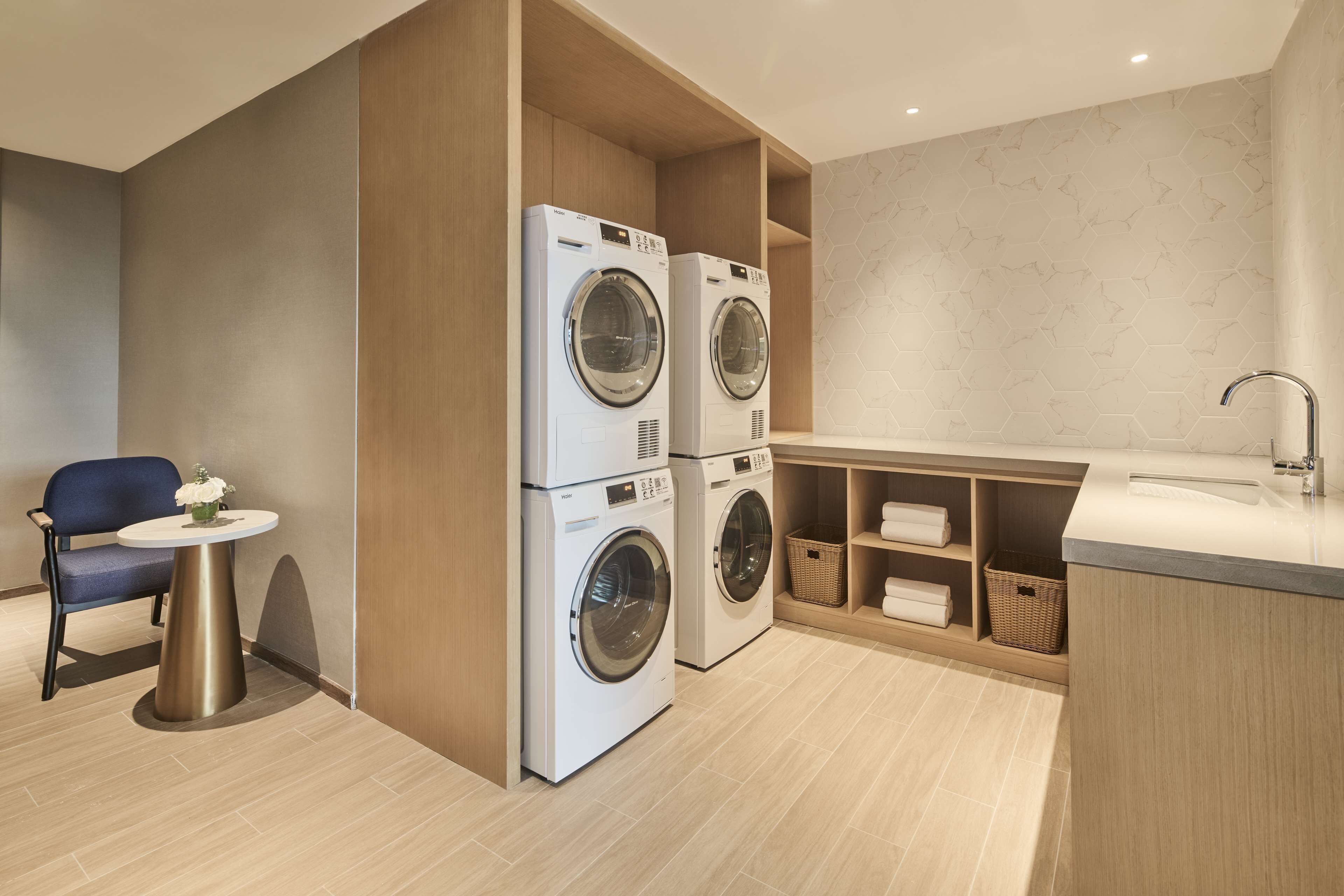 A modern laundry room with two front-loading washing machines, a small round table with a vase, a blue chair, and a built-in sink with shelves for towels.