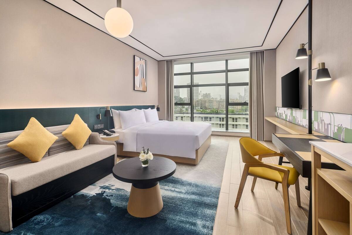 A modern hotel room with a bed, sofa, desk, and a large window offering a city view.