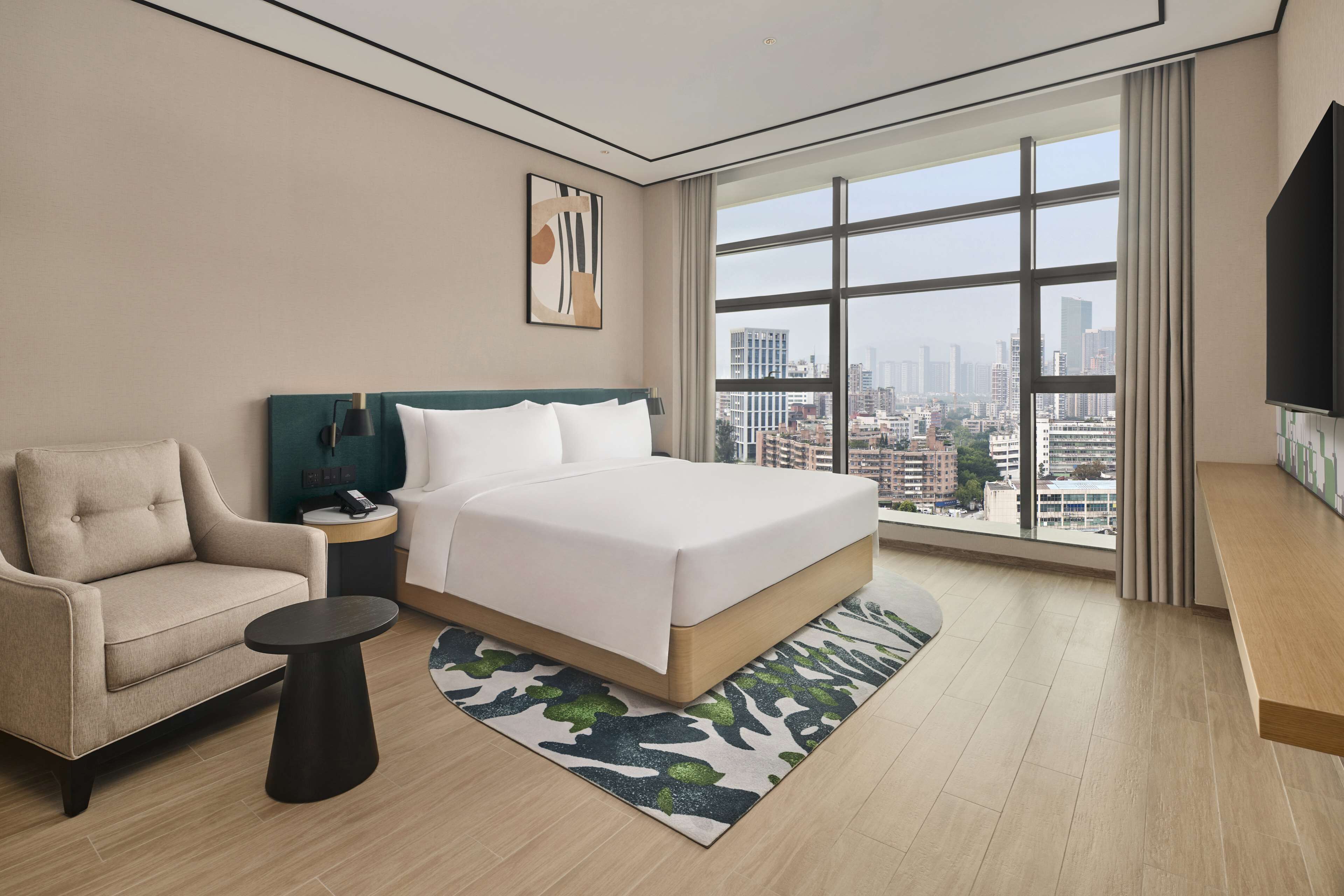 A modern hotel room with a large bed, a sofa, a small table, and a city view through large windows.
