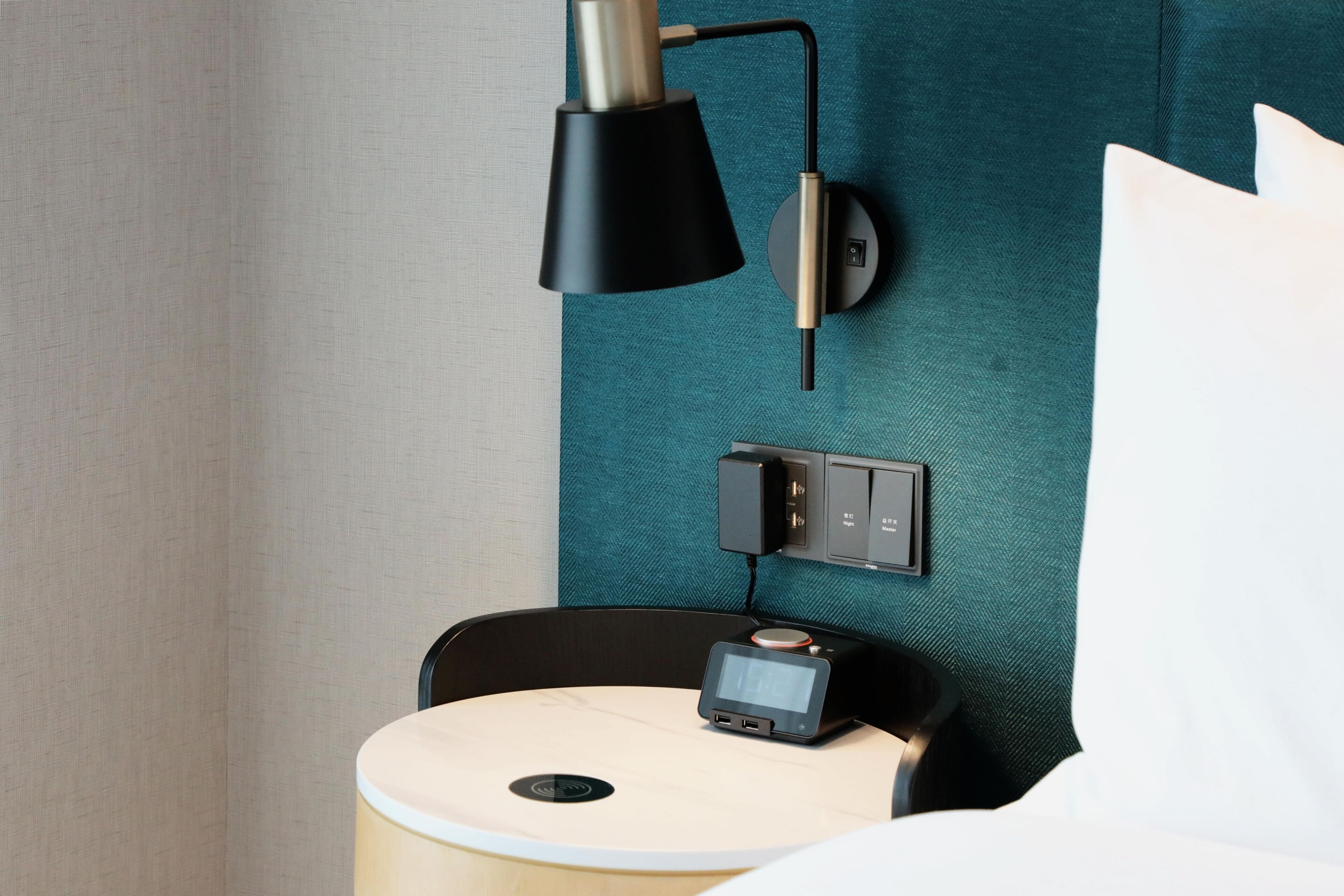 Une chambre d’hôtel moderne avec un mur texturé de couleur turquoise, une lampe noire, une petite table de chevet dotée d’une horloge numérique et une prise électrique.