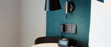 Une chambre d’hôtel moderne avec un mur texturé de couleur turquoise, une lampe noire, une petite table de chevet dotée d’une horloge numérique et une prise électrique.