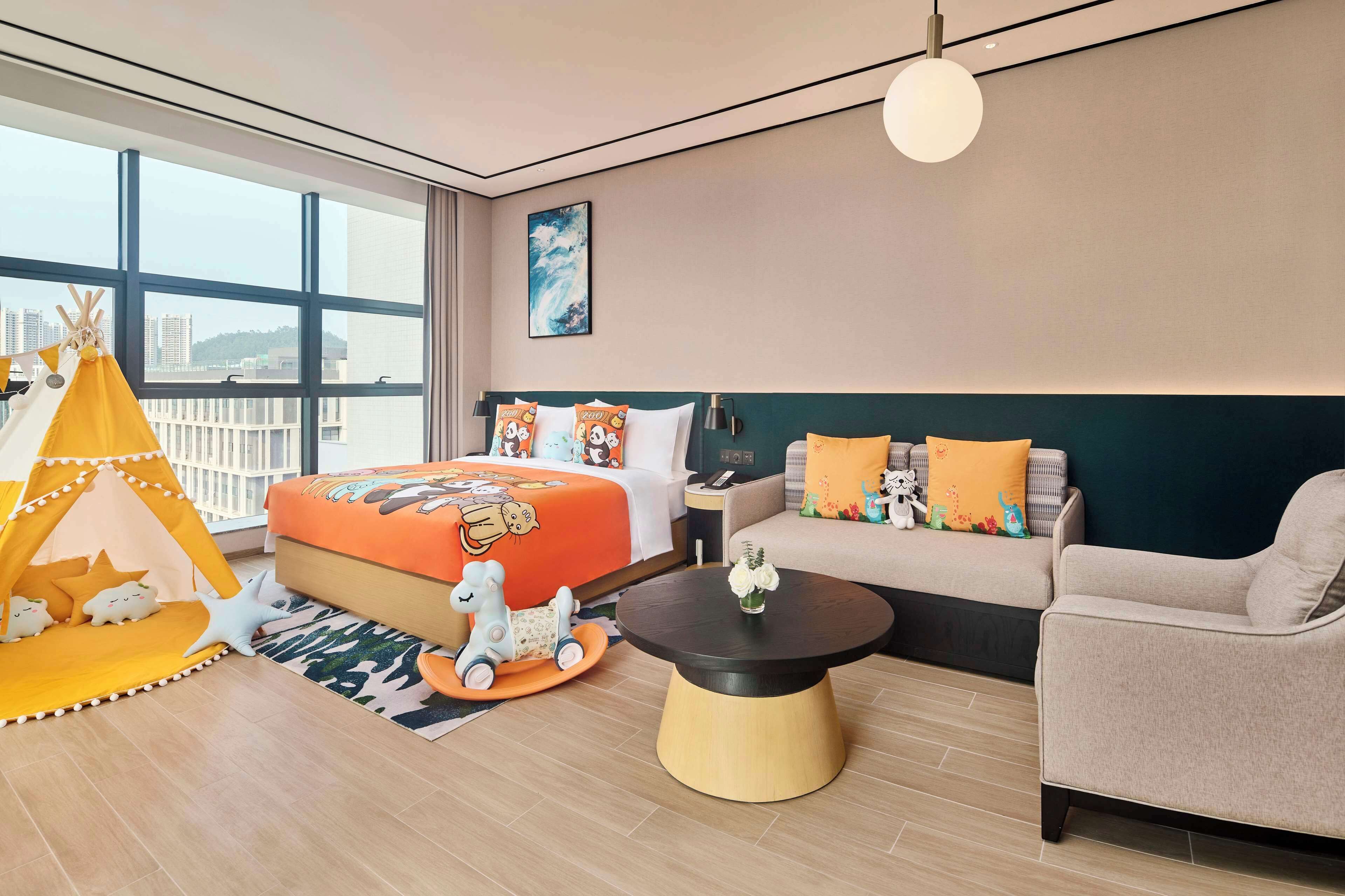 A modern hotel room with a large bed, a sofa, a coffee table, and a play area including a tent and toys.