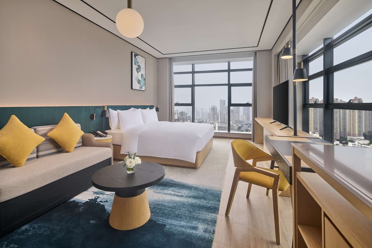 A modern hotel room with a large bed, a sofa, a desk, and a view of the city.