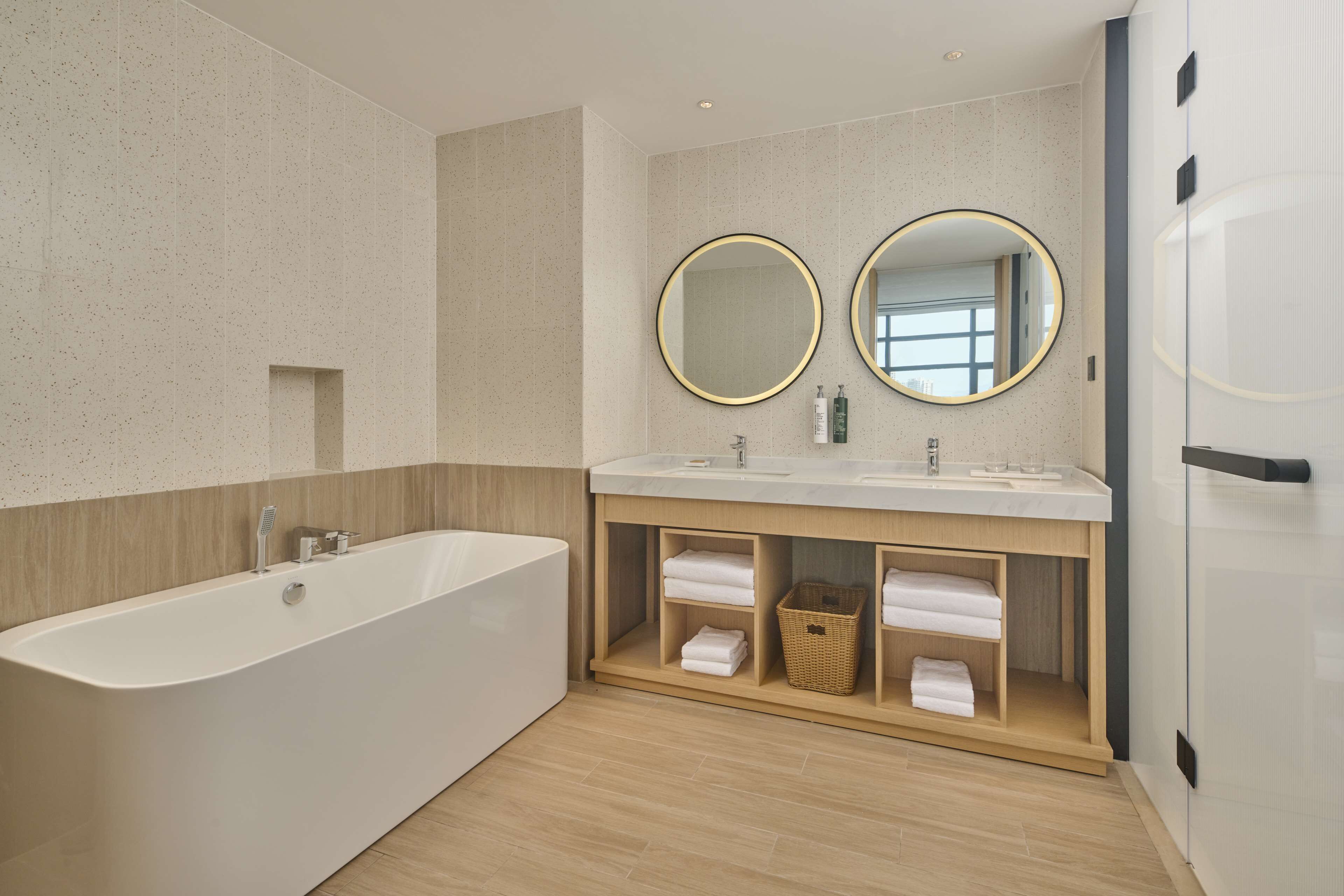 A modern bathroom with a freestanding bathtub, two round mirrors, and a wooden vanity with built-in shelves.