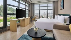 Una habitación de hotel moderna con una cama grande, un escritorio con televisor de pantalla plana, una mesa de centro redonda y vistas a la ciudad a través de amplias ventanas.