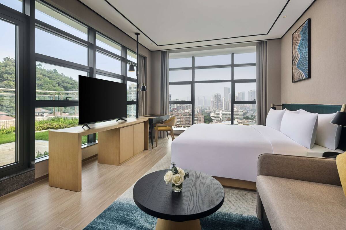 A modern hotel room with a large bed, a desk with a flat-screen TV, a round coffee table, and a view of the cityscape through large windows.