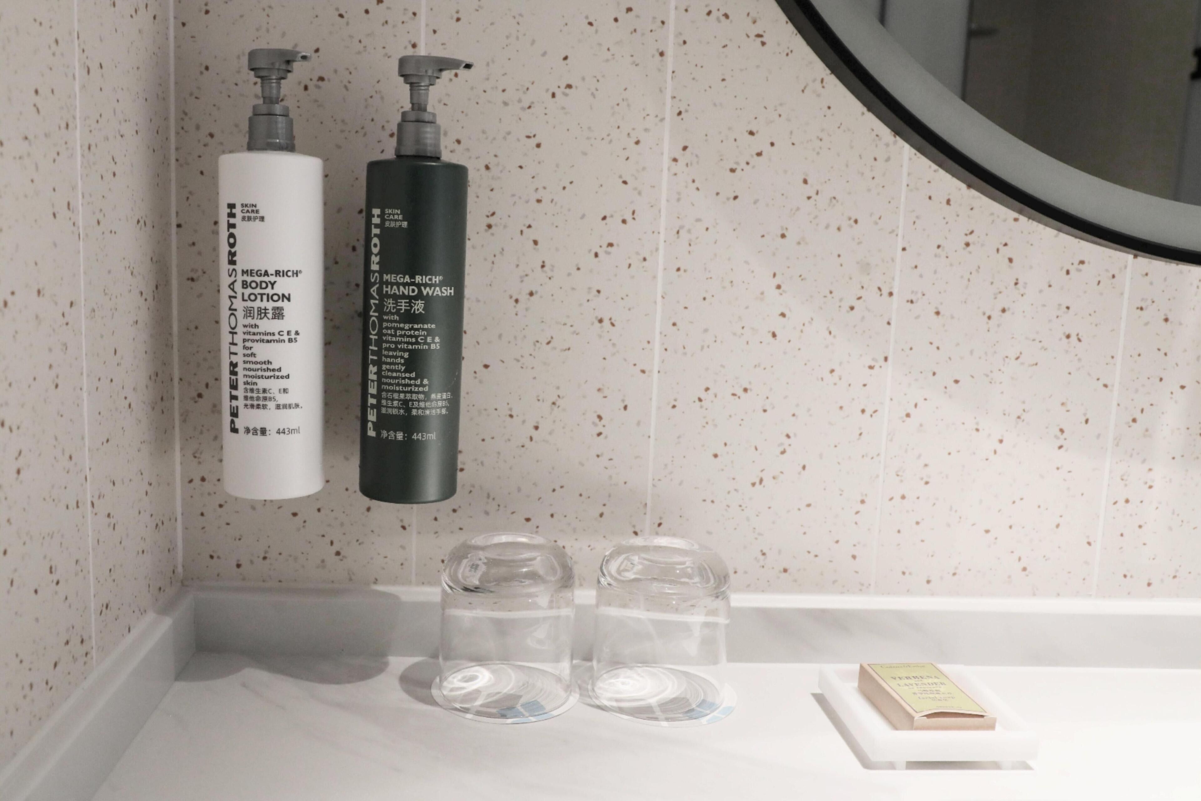A bathroom with two bottles of Mega-Rich body lotion and hand wash, two clear glasses, and a small book on the counter.