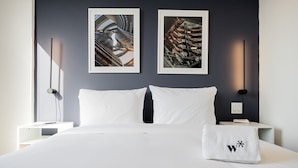 Une chambre d’hôtel avec un grand lit, deux œuvres d’art encadrées au mur, une table de chevet avec une lampe et une serviette ornée d’un logo.
