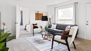 En moderne stue med en grå sofa, en hvid lænestol og et sofabord. Der er et stort vindue med gardiner, en gulvlampe og et mønstret tæppe.