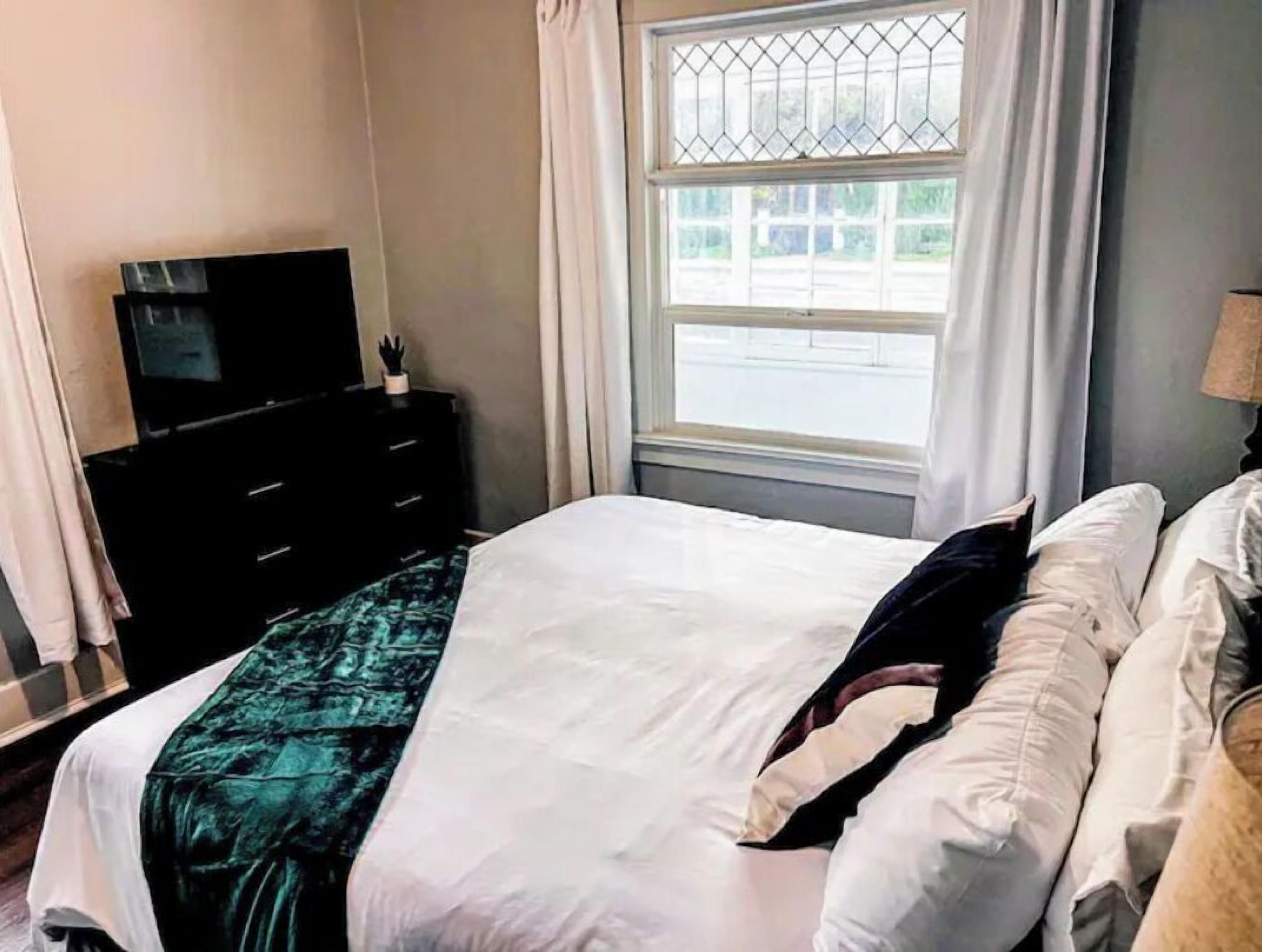 Une chambre à coucher avec un lit, une commode, un téléviseur et une fenêtre avec des rideaux.
