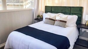 A neatly made bed with white and dark blue bedding, a green upholstered headboard, and a small bedside table with a lamp and a plant. - Modern Comfort in a Bright Nampa Retreat (Nampa)