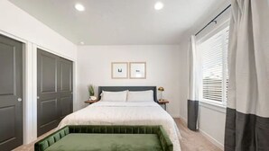 A bedroom with a bed, a green bench, a nightstand, a potted plant, and a window with curtains. - New Modern Cozy Home in Meridian (Meridian)