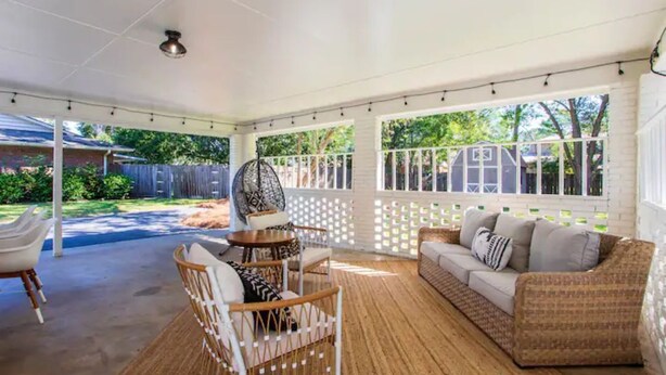Un patio cubierto con techo blanco, suelo de madera y un área de descanso con sofá y sillones.