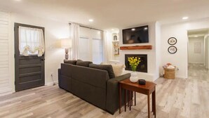 Una sala de estar moderna con un sofá de color oscuro, una pequeña mesa de madera con un jarrón de flores, una chimenea y un televisor de pantalla plana montado en la pared.