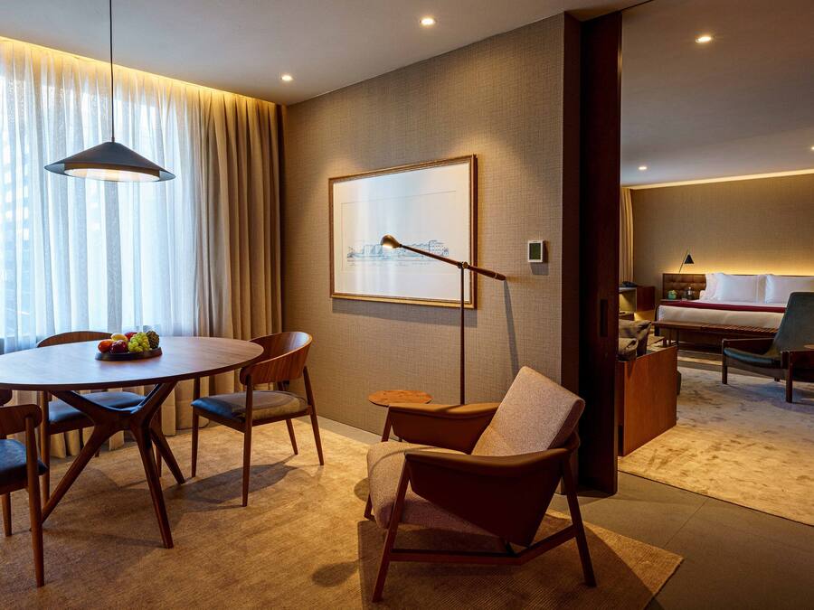 Quarto de hotel moderno com área de refeições, ampla janela com cortinas leves, obra de arte com vista urbana emoldurada, espaço de estar aconchegante com sofá e poltronas, e quarto separado com cama e mesas de cabeceira.