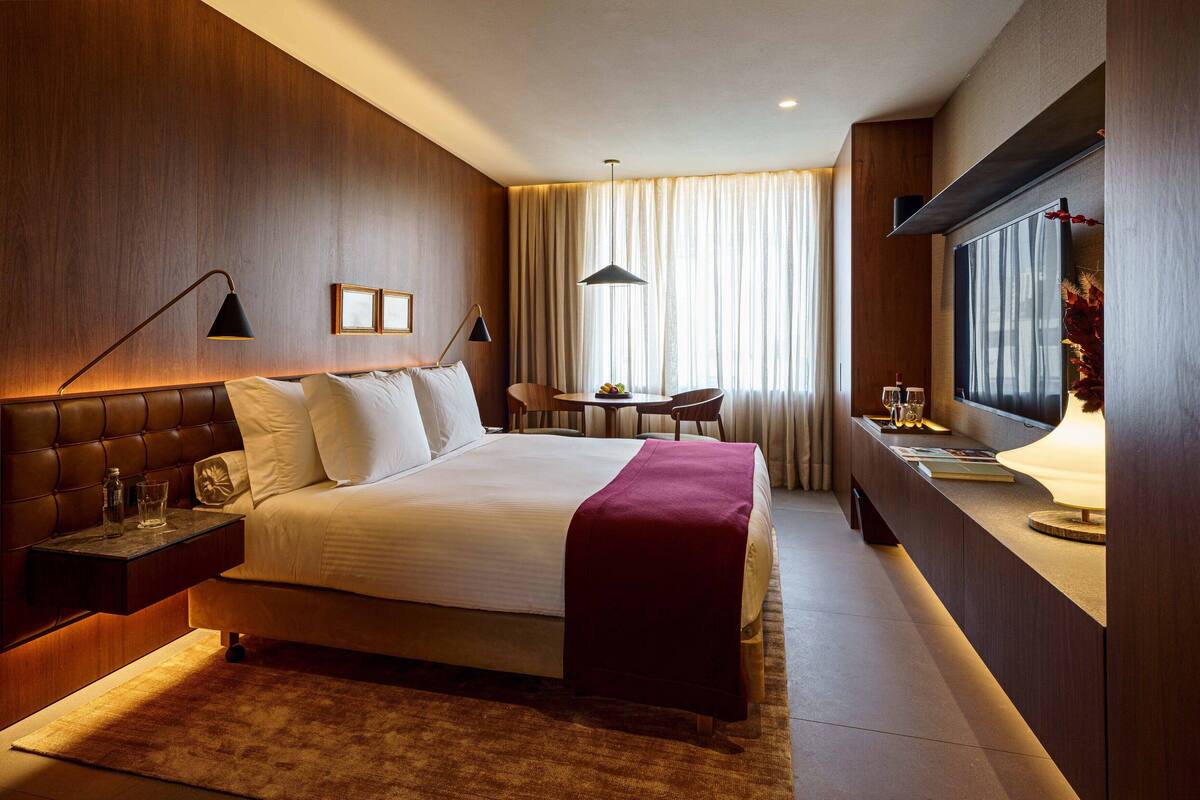 A hotel room with a large bed, bedside tables, a desk with a lamp, a chair, a small table with fruit and glasses, and a window with curtains.
