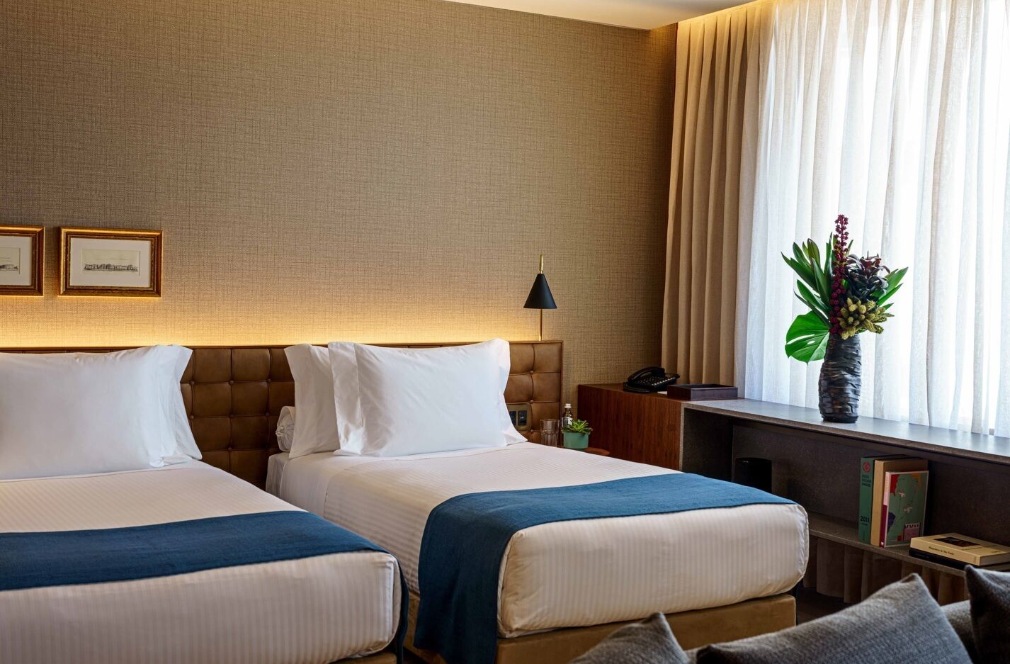 A hotel room with two beds, a desk, a chair, a lamp, and a window with curtains.