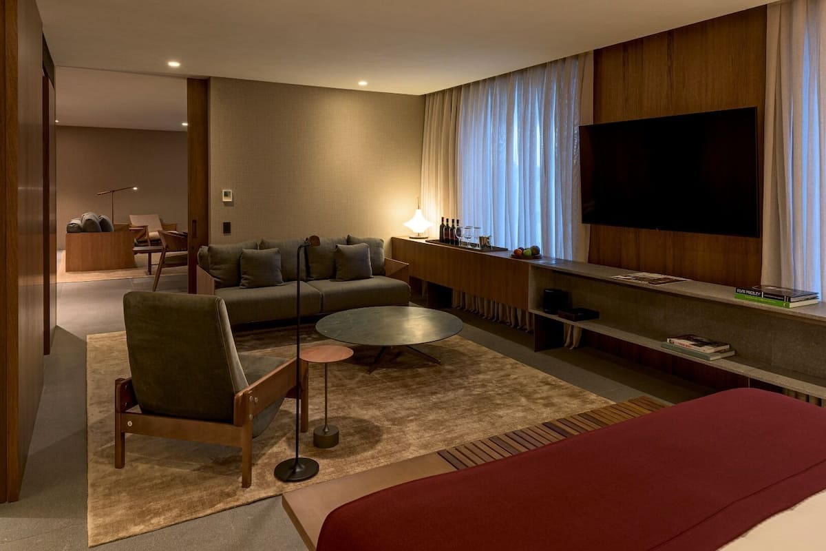 A modern hotel room with a sofa, armchair, round coffee table, and a TV mounted on the wall.