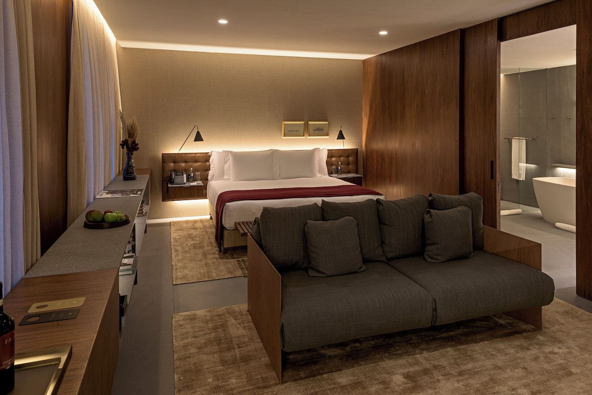 A modern hotel room with a bed, a sofa, a nightstand, a mirror, and a wooden panel wall.