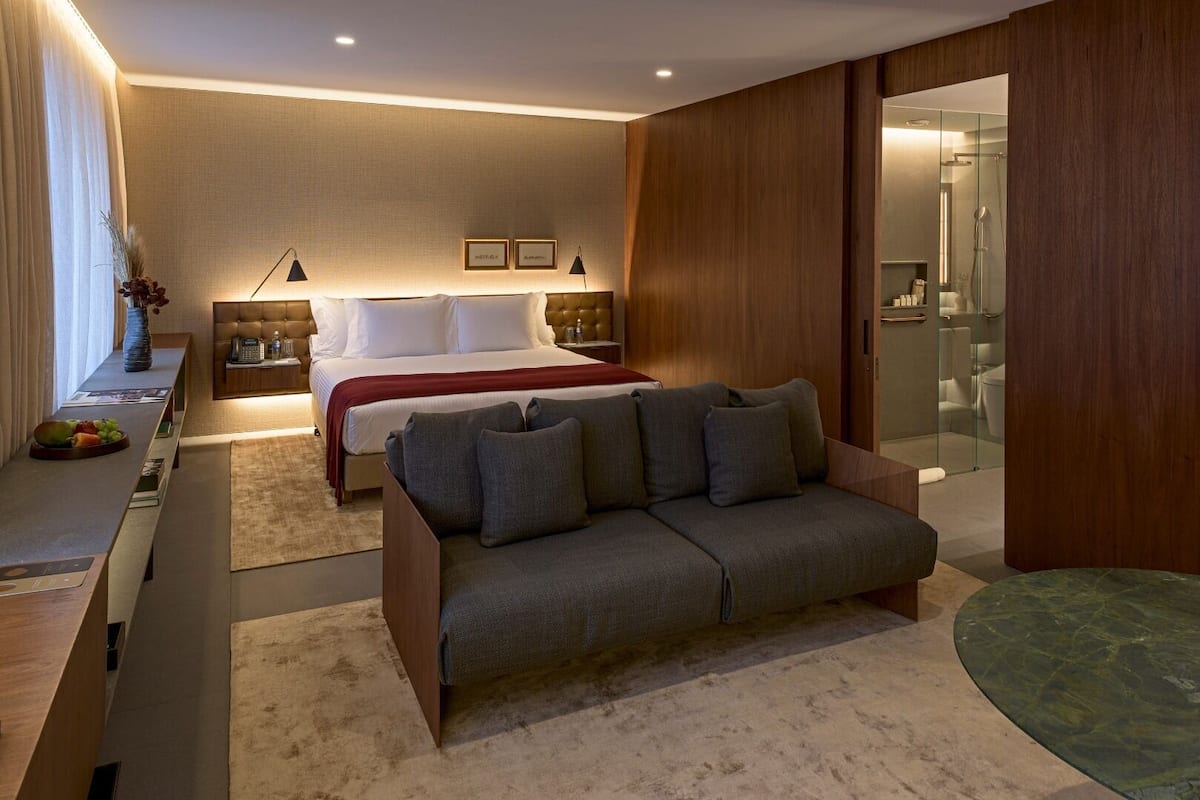 A modern hotel room with a bed, a sofa, a desk, and a glass shower area.