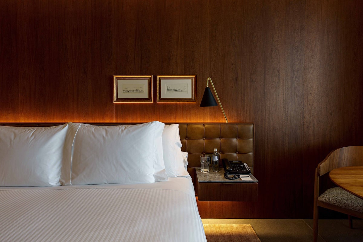 A hotel room with a large bed, a wooden headboard, a bedside table with a lamp, and framed pictures on the wall.