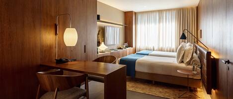 A modern hotel room with a wooden accent wall, a desk with a chair, a bed with a blue blanket, and a bedside table with a lamp.