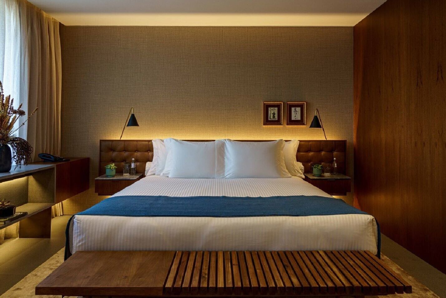 Een hotelkamer met een groot bed, twee nachtkastjes, een houten bankje en een groot raam met gordijnen.