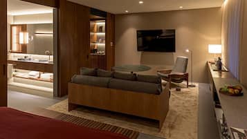 A modern hotel room with a wooden sofa, a small round table, a TV mounted on the wall, and a minibar.