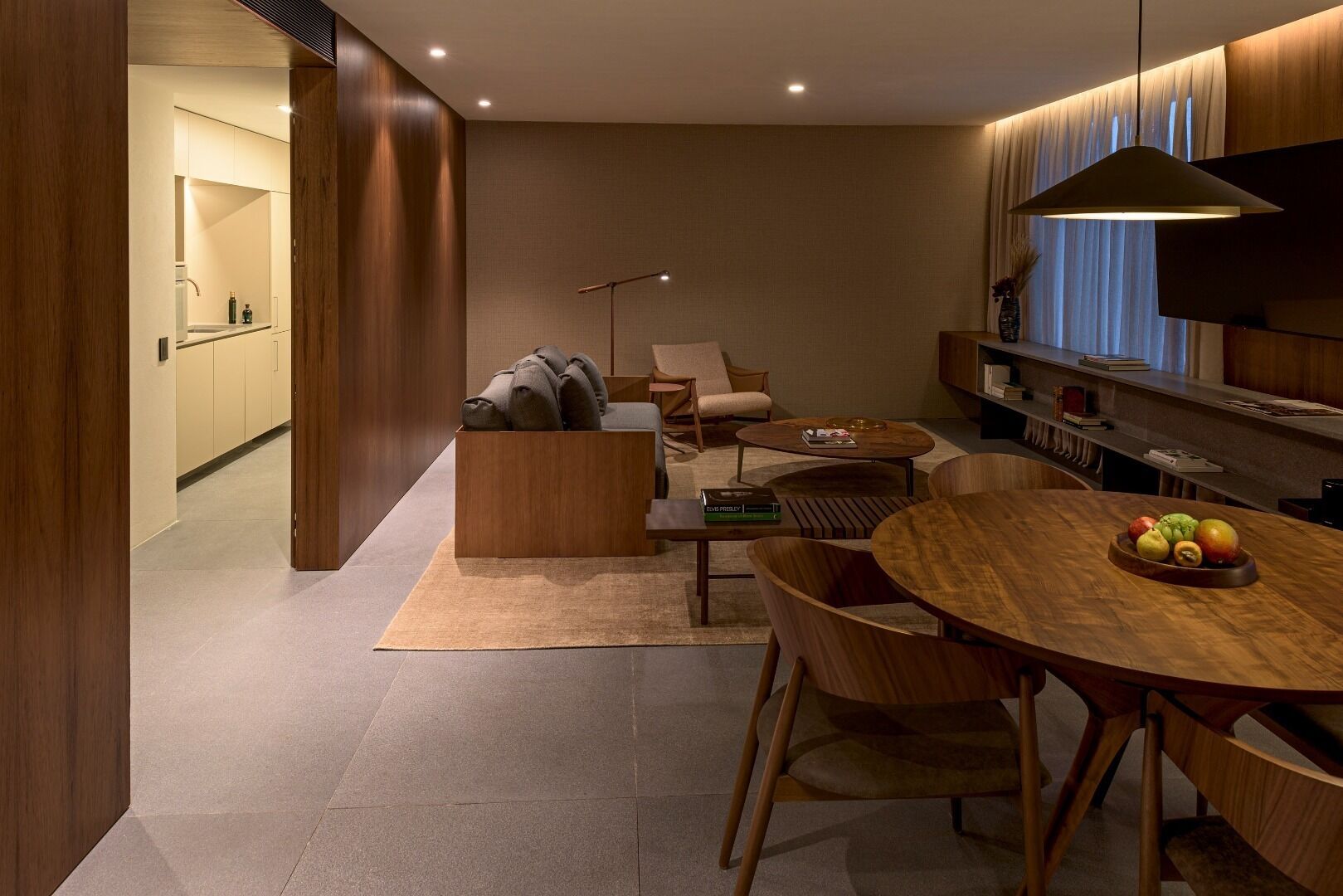 A modern living room with a sofa, coffee table, and dining table. There is a kitchen area in the background with cabinets and a window.