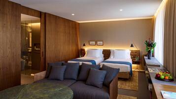 A hotel room with a sofa, two beds, a desk with a fruit bowl, and a bathroom visible through a glass partition.
