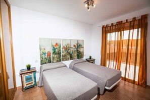 A bedroom with two beds, a painting of parrots, a small table with a plant, and a window with curtains.
