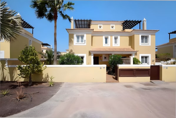 A two-story residential building with a garage, a covered patio, and a small garden with palm trees and shrubs.