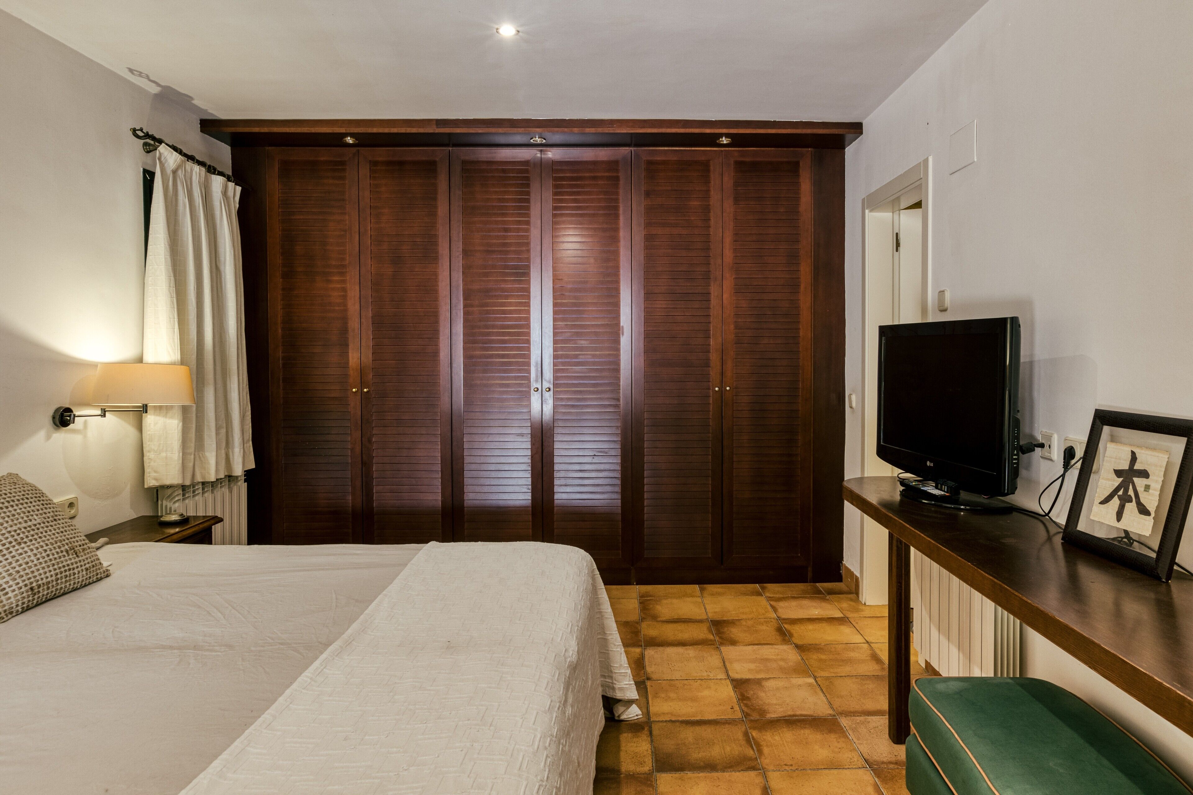 A bedroom with a bed, a TV, a wardrobe, a bedside lamp, and a framed picture on a dresser.