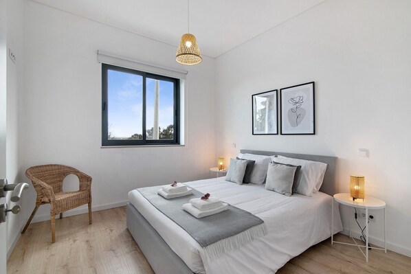 A modern bedroom with a large bed, a wicker chair, a bedside table with a lamp, and a window with a view.