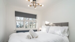 A neatly made bed with white linens and a rolled towel, a grey upholstered headboard, a window with blinds, and a modern chandelier.