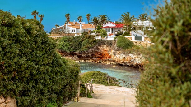 A coastal property with a stone pathway leading to a cliff overlooking the sea, surrounded by lush greenery and palm trees. - Holiday Home 'Aloes' with Shared Pool, Private Terrace and Wi-Fi (Ciutadella de Menorca)