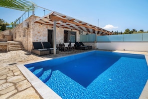 A swimming pool with a stone deck, a covered seating area with chairs, and a staircase leading to an upper level.
