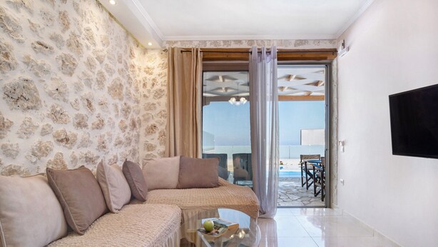 A living room with a stone wall, a sofa set, a glass coffee table, and a view through a glass door. - Holiday Apartment "Suite Irida" with Sea View, 2 Terraces & Wi-Fi (Lefkada)